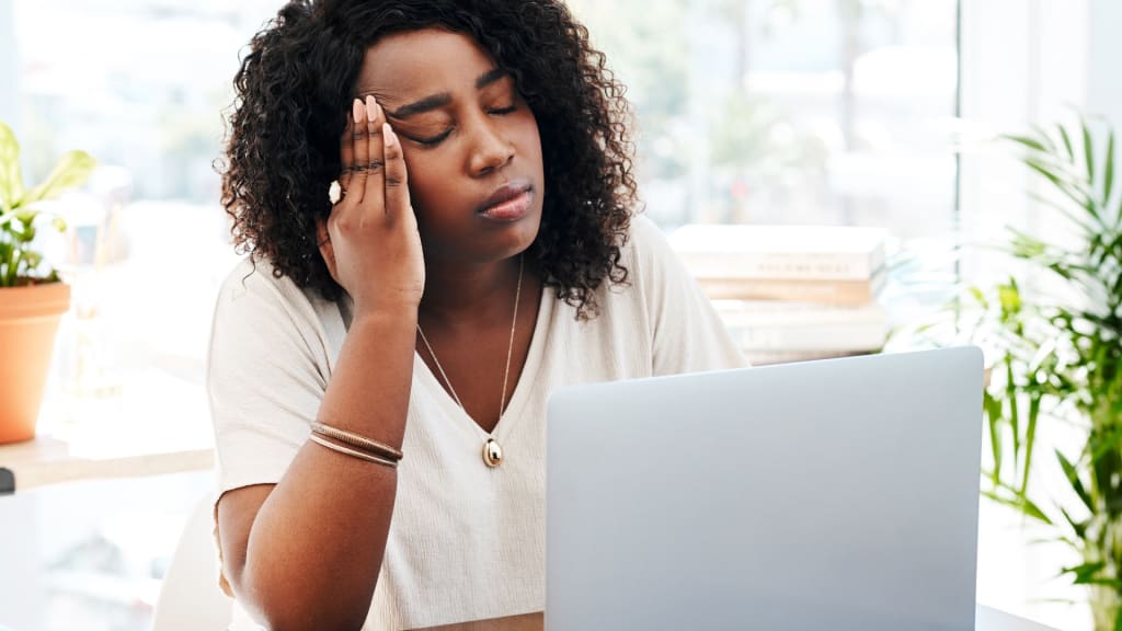 A woman sits in front of a laptop with her eyes closed, touching her temple as if experiencing a headache or stress.